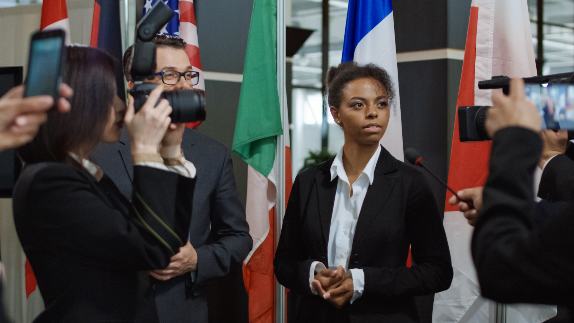 A woman in a suit talks to journalists in front of flags