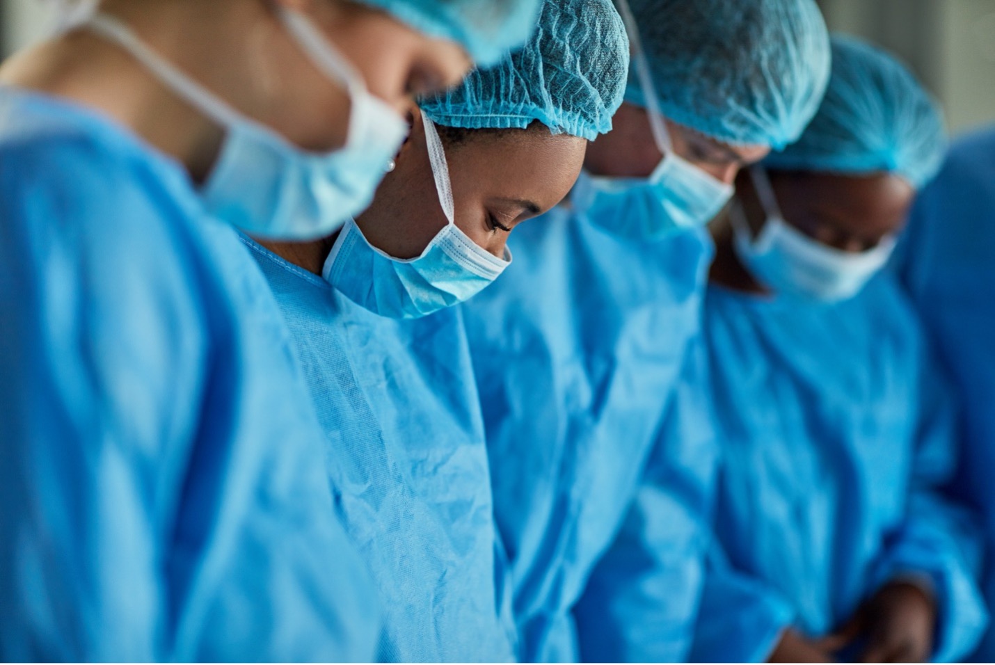 several dark-skinned medical professionals in blue scrubs, masks, and hairnets with heads bowed